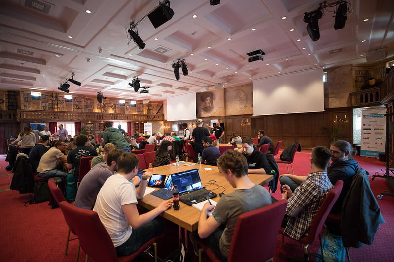 A bunch of people sat around tables. They're all hacking on laptops.